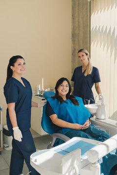 Portrait of three women, dentist, assistant and patient in a dental office smiling and looking at camera.