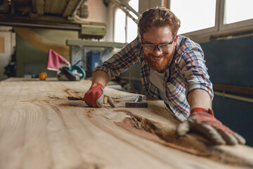 Close up of smiling craftsman working with chisel while cutting wooden plank in carpentry workshop - obrazy, fototapety, plakaty
