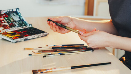 Art tools. Painting accessories. Unrecognizable woman artist hands with paintbrushes watercolor paint palette at artwork table studio workplace.