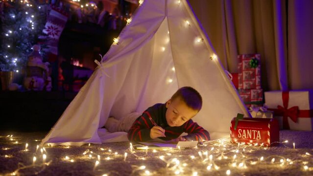 Preschooler boy lying near decorated Christmas tree at fireplace and writing letter with his wish list and big dreams to Santa Claus to send through red mailbox to Santa Village in North Pole slowmo