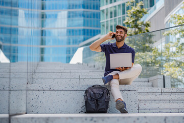 Male freelancer sitting with laptop on city skyscrapers background and talking phone 