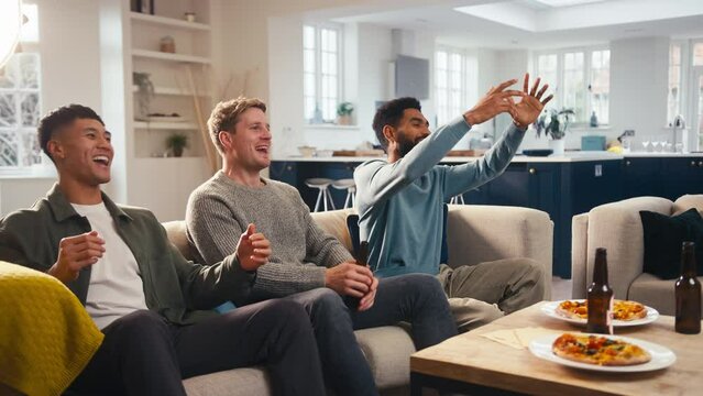 Group of excited male friends at home in lounge watching sports on TV celebrating with beer and pizza together - shot in slow motion