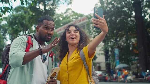 Medium shot of young diverse couple taking selfies on smartphone in street in Vietnam, biracial woman holding out gadget and smiling, and Indian man showing V-sign and making funny faces