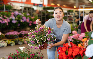 Cheerful young girl choosing potted colorful blooming fuchsia in flower shop to decorate courtyard...