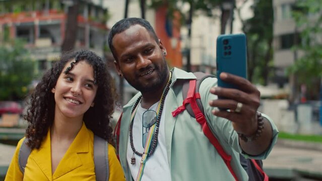 Medium close-up shot of ethnically diverse couple with backpacks standing in city street in South East Asia while sightseeing on holiday, posing, smiling and taking selfie together on smartphone
