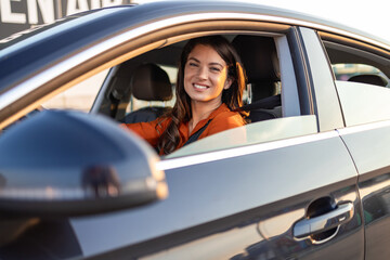 Beautiful young happy smiling woman driving her new car at sunset. Portrait of smiling young Caucasian woman sitting alone on driver seat with fasten belt while driving modern car.