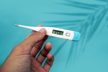 A man's hand holding a digital thermometer in her hand on a blue background with leaves shadow. A person looks at an electronic thermometer with temperature.