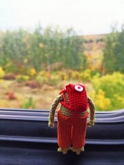 A toy frog with a red backpack looks out the window on a train.  There are autumn trees outside the window.