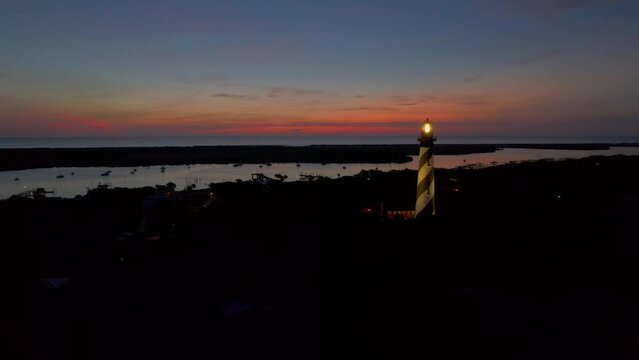 Painted sky backdrop with boats along the coastal inlet in Saint Augustine Florida in the early morning. A lighthouse Illuminated and spinning its lantern around the coast.