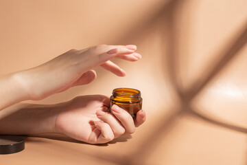Female applying a collagen firming cream to a hand. Open cream jar on beige background. Cosmetics mockup. Trendy colors and shadows.