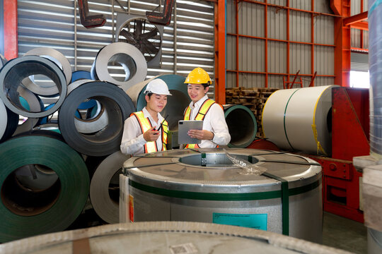 Industrial Warehouse Scene Featuring Two Safety-conscious Men Inspecting Steel Sheet Roll Stack, Imbued With The Raw Essence Of Daily Manual Labor And Meticulous Attention To Detail.