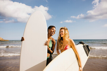 Two cheerful female friends celebrating during a day off at the beach doing surf. Happy women in swimwear holding their surfboards on the shore, teaming up for a journey of surfing.