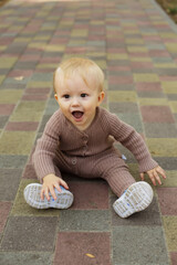 Fototapeta premium a little boy in a brown jumpsuit sits happily on the sidewalk in the park