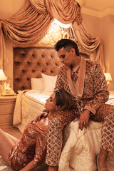 Fototapeta premium Wealthy young couple is dressed in radiant golden outfits adorned with sequin embroidery, situated in a luxurious suite