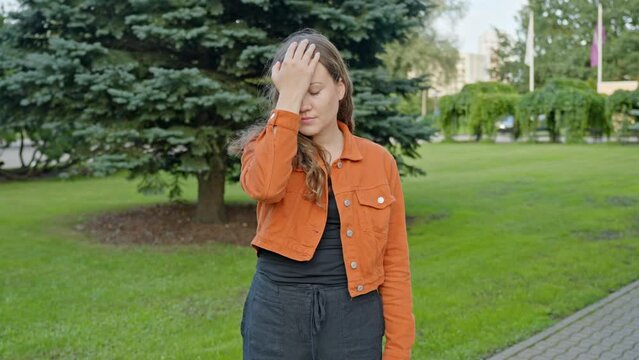 Young Woman Doing Palm Face in a City Park with a Forgetful Emotion Wearing an Orange Jacket.