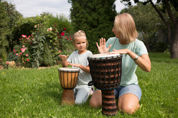 mom and child daughter play at ethnic drums sitting on the grass on a sunny day. a family hobby. child learn to play drums