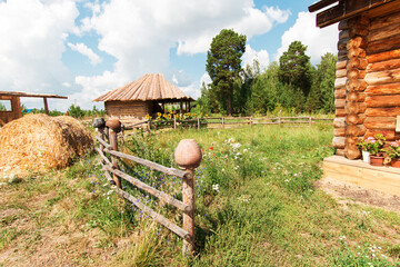 Village yard with buildings. Clay pots hang on the fence. Yellow hay. Wildflowers in a green meadow. Blue sky with white clouds. There is a forest in the background.