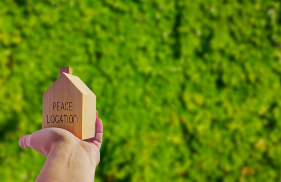 Wooden house with 'freedom location' written on it and a human hand holding it. Green background. Photograph.