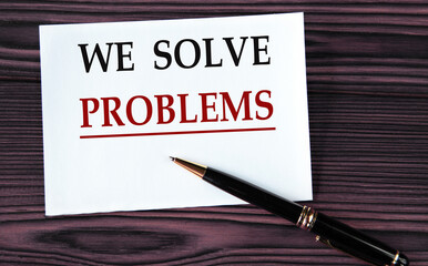 WE SOLVE PROBLEMS - words on a white sheet on a brown wooden background with a pen