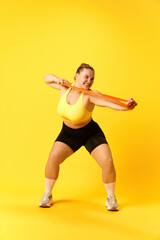 Fototapeta na wymiar Full length portrait of confident plus size sports woman in tracksuit training hard with sport elastic bands for hands over yellow background.