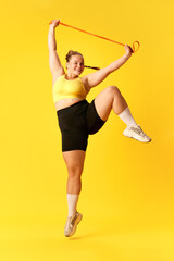 Fototapeta na wymiar Full length portrait of plus size woman dressed sporty training, jumping with sport elastic band against over yellow background.