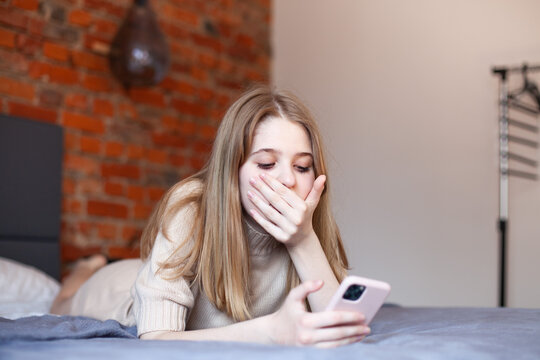 Surprised teen girl lying on bed looking at smartphone covering mouth with hand. Teens and gadgets, online courses for teens, online shopping, breaking news. Selective focus.