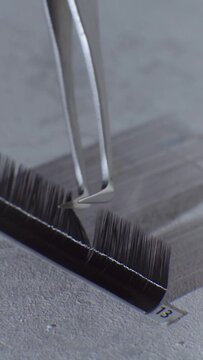Video of tweezers in hand of unrecognizable lashmaker running tip of tool over black false eyelashes in palette. Professional eyelash extension pincette perfect for picking up tiny cilia. Vertical.
