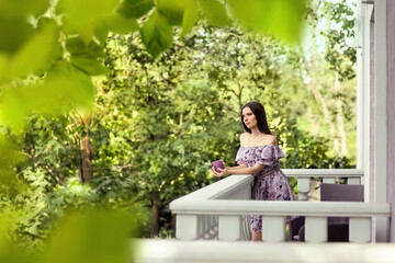 A girl in a lilac dress stands on the balcony of her house. She is thoughtful, holding a mug of tea in her hands. Season of the year is summer.