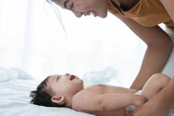 Asian mother playing in bed with her 3 month old newborn baby while changing diaper on her little daughter, look at each other, smile and laugh together
