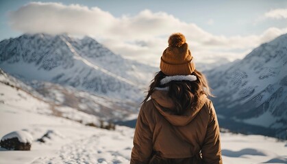 Fototapeta na wymiar Rear view of a young traveler girl in a wool hat yellow shirt standing over snow-covered mountain peaks. Winter travel scene, wanderlust concept