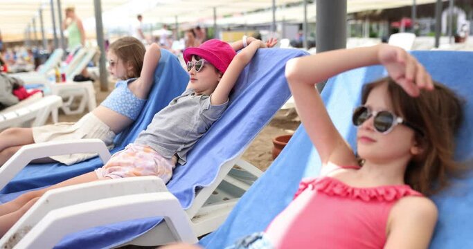 Three happy girls lie on sun loungers and look at sea. Children on beach on sunny day during summer vacation