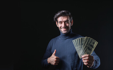 Portrait of a happy young businessman in a sweater displaying cash and giving the thumbs up, isolated on a black background with copy space for content.