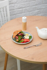 Breakfast of croissant burger with fries ham, boiled eggs with teapot on round wooden table on light bricks background.