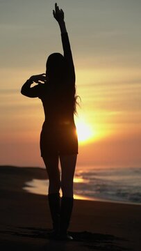 Rear view silhouette woman with hands raised standing on sandy beach at sunrise. Female in summer short dress, high boots during weekend beach holiday at golden hour. Slow motion, handheld, vertical