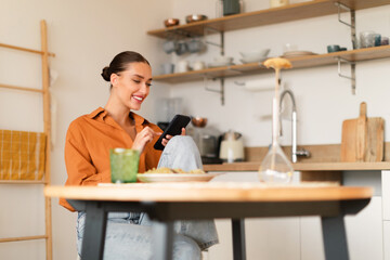 Positive european lady using cellphone while having lunch, sitting at table in kitchen, free space