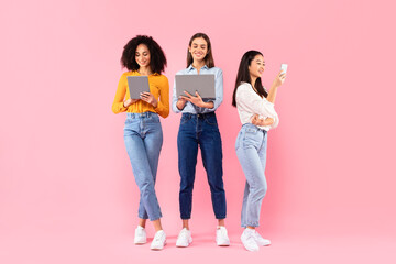 Happy multiracial ladies friends using different gadgets, using smartphone, laptop and digital tablet, pink background