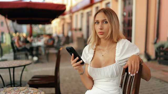 Close-up portrait of a beautiful young blonde woman with charming eyes holding a smartphone in her hand, sitting at a table in a street cafe and smiling sweetly outdoors