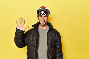 Man in winter coat with snow goggles on yellow smiling cheerful showing number five with fingers.