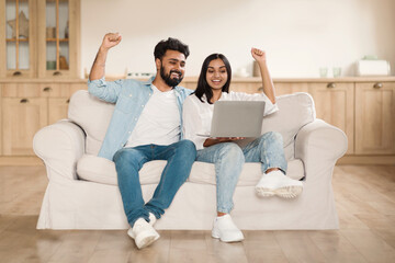 Giveaway, lottery, cashback. Overjoyed indian couple using laptop sitting on couch at home, gesturing and exclaiming