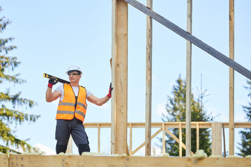 Side view of man wearing helmet and uniform standing on wooden concstrction, holding measuring tool. Builder, worker building house in forest in summer. Concept of building.