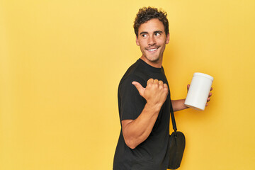 Young Latino with protein bottle on yellow points with thumb finger away, laughing and carefree.