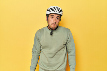 Casual dressed Latino cyclist on yellow shrugs shoulders and open eyes confused.