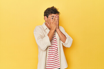 Young Latino man posing on yellow background blink through fingers frightened and nervous.