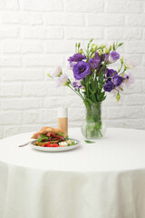 Obraz na płótnie Canvas Breakfast of croissant burger with fries ham, boiled eggs on round white table with vase of bouquet of eustomas on light bricks background.