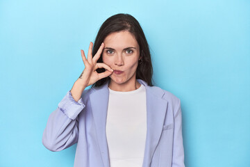 Woman in blue blazer on blue background with fingers on lips keeping a secret.