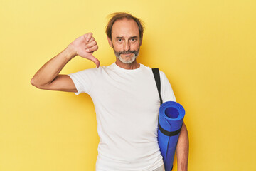 Man with yoga mat in yellow studio showing a dislike gesture, thumbs down. Disagreement concept.