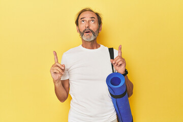 Man with yoga mat in yellow studio pointing upside with opened mouth.