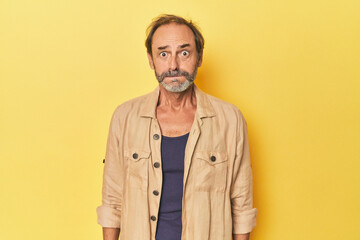 Caucasian middle-aged man in yellow studio blows cheeks, has tired expression. Facial expression concept.