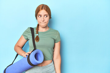 Redhead young woman holding yoga mat in studio confused, feels doubtful and unsure.
