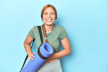 Redhead young woman holding yoga mat in studio laughing and having fun.